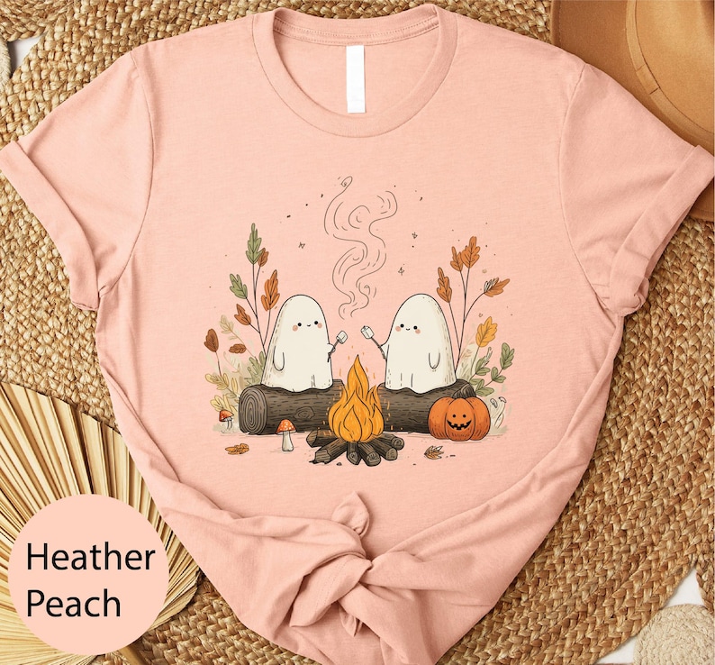 A pink t-shirt with a whimsical graphic design depicting two ghostly figures sitting by a campfire, surrounded by autumn foliage and pumpkins.