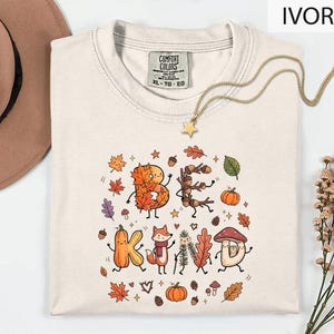 May include: An ivory short-sleeve t-shirt with a graphic print featuring the text BE KIND formed by autumn-themed characters like leaves, acorns, a pumpkin, a fox, and a mushroom, surrounded by small seasonal icons like stars and pinecones.