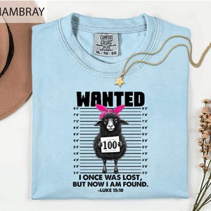 May include: A light blue t-shirt featuring a graphic of a black sheep wearing a pink headband, standing against a height chart. The text reads WANTED, 100, and I ONCE WAS LOST, BUT NOW I AM FOUND. -LUKE 15:10. The shirt has a Comfort Colors label.