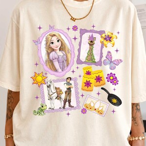May include: An ivory t-shirt featuring a graphic print of Rapunzel, a tower, a horse, a chameleon, a frying pan, lanterns, a crown, and flowers. The text IVORY is printed in the top left corner. The design includes purple frames and sparkles.