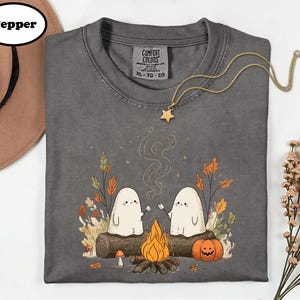 A gray t-shirt with a Halloween-themed graphic design, including two ghostly figures, a campfire, and autumn foliage. The t-shirt is accompanied by a hat, a necklace, and some flowers.