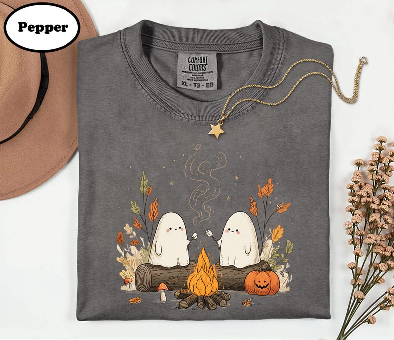 A gray t-shirt with a Halloween-themed graphic design, including two ghostly figures, a campfire, and autumn foliage. The t-shirt is accompanied by a hat, a necklace, and some flowers.