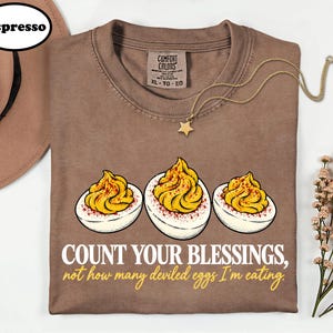 May include: An espresso-colored t-shirt featuring a graphic of three deviled eggs with yellow yolks and red paprika seasoning. The text reads: COUNT YOUR BLESSINGS, not how many deviled eggs I'm eating. The shirt has a Comfort Colors label.