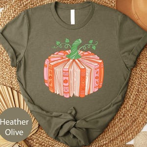 A t-shirt with a graphic design of a pumpkin made up of books, placed on a woven mat.