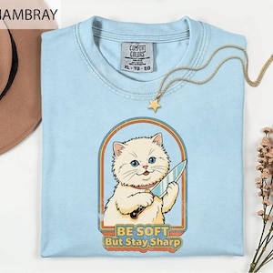 May include: Light blue t-shirt with a graphic of a white cat holding a knife, framed by a retro design. The text "BE SOFT But Stay Sharp" is below the cat. A gold necklace with a star pendant is on the shirt.