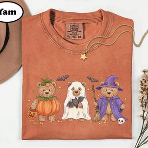 May include: A rust-colored t-shirt featuring a graphic of three teddy bears in Halloween costumes: one as a pumpkin, one as a ghost holding a bat, and one as a wizard with a broom and hat. The shirt has a Comfort Colors label in size XL.