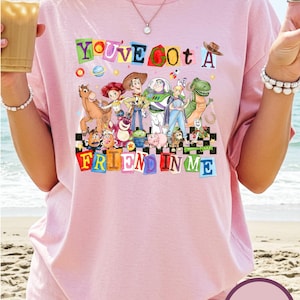 A woman wearing a pink t-shirt with a colorful graphic of characters from Toy Story, including Woody, Buzz Lightyear, and other beloved characters, along with the text "You've got a friend in me".