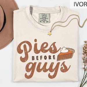 May include: An ivory crew neck t-shirt with a Comfort Colors label, featuring the brown text 'Pies before guys' and a graphic of a slice of pumpkin pie with whipped cream. A gold-toned star pendant necklace rests on the neckline of the shirt.