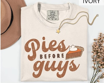 May include: An ivory crew neck t-shirt with a Comfort Colors label, featuring the brown text 'Pies before guys' and a graphic of a slice of pumpkin pie with whipped cream. A gold-toned star pendant necklace rests on the neckline of the shirt.