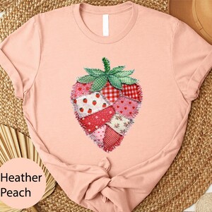 May include: Heather peach-colored t-shirt featuring a patchwork strawberry design. The strawberry is composed of various fabric patterns in red, pink, and white, with a green leaf crown. The shirt's color is labeled "Heather Peach".