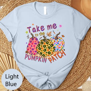A light blue t-shirt with a colorful graphic design featuring three spotted pumpkins and the text "Take me to the pumpkin patch" printed on it.