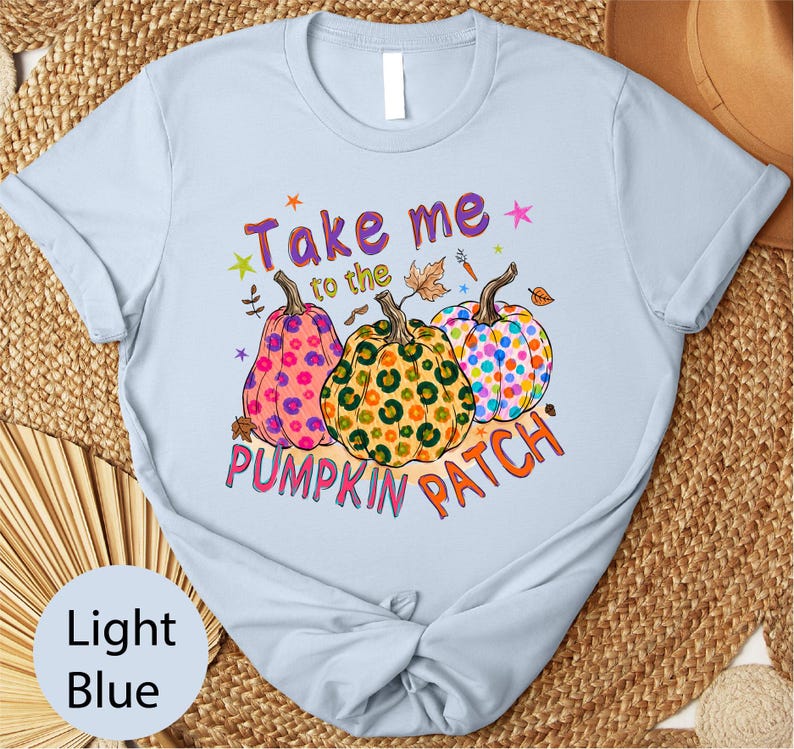 A light blue t-shirt with a colorful graphic design featuring three spotted pumpkins and the text "Take me to the pumpkin patch" printed on it.