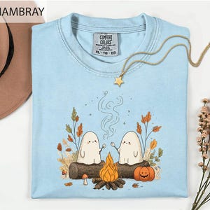 a light blue t-shirt with a whimsical graphic design depicting two ghostly figures roasting marshmallows over a campfire, surrounded by autumn foliage and a star.