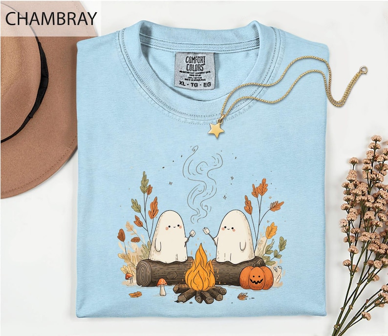 a light blue t-shirt with a whimsical graphic design depicting two ghostly figures roasting marshmallows over a campfire, surrounded by autumn foliage and a star.