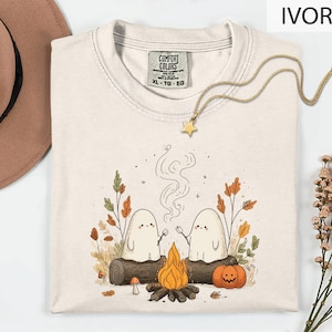 a white t-shirt with a graphic design depicting two ghostly figures roasting marshmallows over a campfire, surrounded by autumn foliage and a pumpkin.