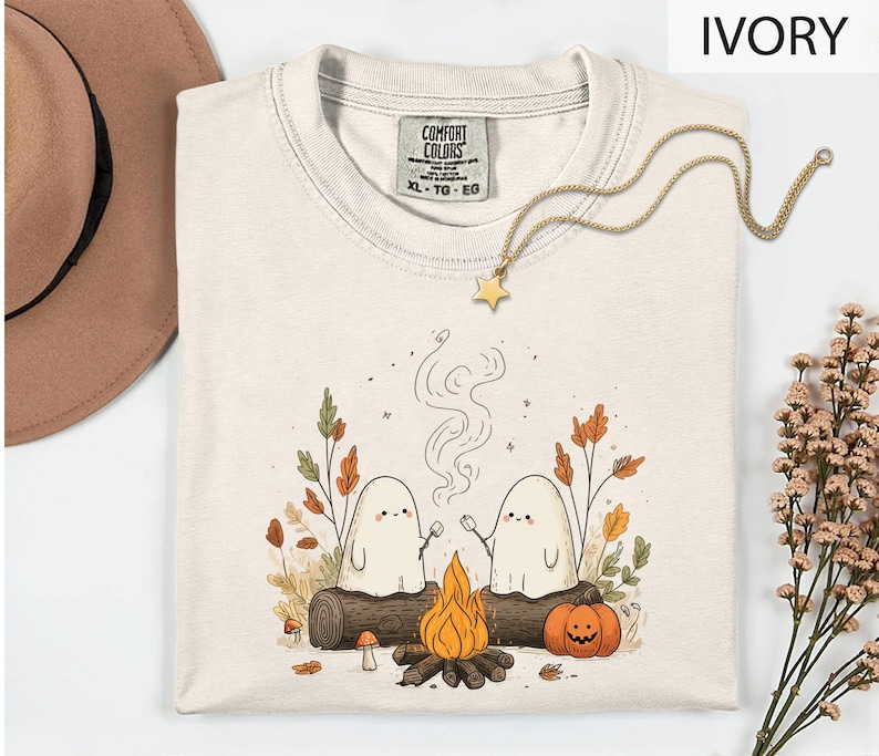 a white t-shirt with a graphic design depicting two ghostly figures roasting marshmallows over a campfire, surrounded by autumn foliage and a pumpkin.