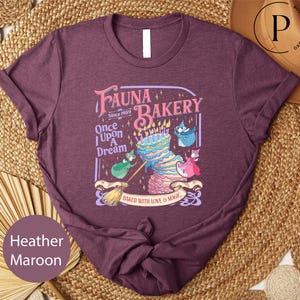 May include: Heather maroon t-shirt with a whimsical "Fauna Bakery" graphic. The design features a layered cake with candles, characters, and the text "Baked with Love & Magic."