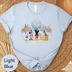 A light blue t-shirt with a graphic design featuring Disney characters Mickey Mouse, Minnie Mouse, Donald Duck, and Goofy standing in front of the Earth's Fair Park attraction.