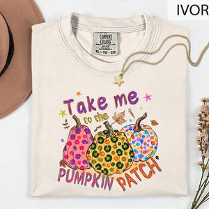 a white t-shirt with a colorful graphic design of three pumpkins and the text "Take me to the pumpkin patch" printed on it. The t-shirt is laid out on a white surface, with a hat and some flowers visible in the background.