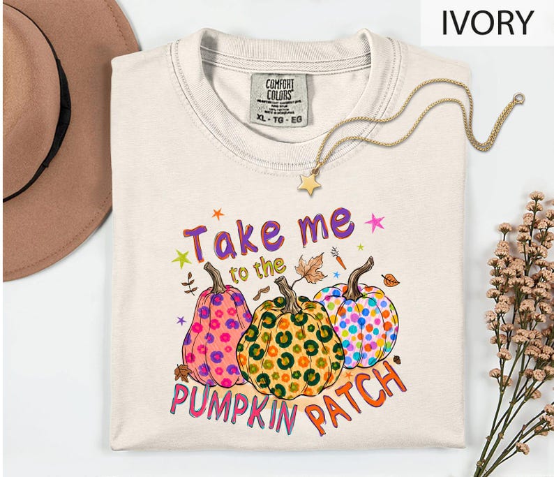 a white t-shirt with a colorful graphic design of three pumpkins and the text "Take me to the pumpkin patch" printed on it. The t-shirt is laid out on a white surface, with a hat and some flowers visible in the background.