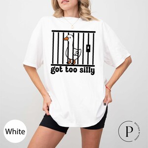 Peut inclure: T-shirt blanc avec un motif noir d'un canard de dessin anim&eacute; derri&egrave;re des barreaux, avec le texte "got too silly" en noir. Le canard a un bec et des pattes oranges. Le t-shirt est de style d&eacute;contract&eacute;.