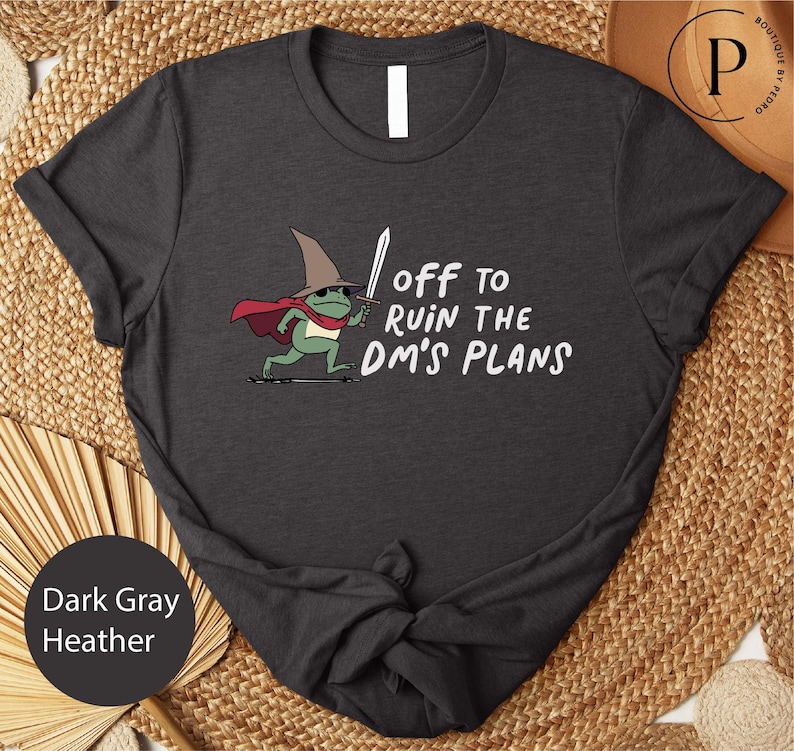 A black t-shirt with a graphic design depicting a frog wearing a wizard's hat and holding a sword, along with the text "Off to ruin the dm's plans".