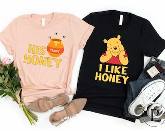 May include: Two t-shirts, one pink and one black, with a Winnie the Pooh theme. The pink shirt says "His Honey" with a honey pot and bees. The black shirt says "I Like Honey" with a picture of Winnie the Pooh.