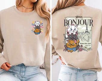 May include: A beige sweatshirt with a graphic print of three cats from the Disney movie The Aristocats. The print includes the text "Bonjour" and a depiction of the Eiffel Tower. The sweatshirt has a crew neck and long sleeves.