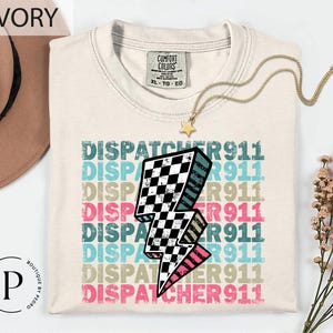 May include: Ivory-colored t-shirt with the text "DISPATCHER 911" in multiple colors and a checkered lightning bolt graphic. The shirt has a crew neck and a Comfort Colors tag. A gold necklace with a star pendant is draped on the shirt.