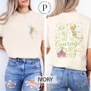 May include: Ivory-colored t-shirt featuring a cartoon fairy with a wand on the front. The back of the shirt displays the text "We ALL HAVE THE Courage TO FLY" with a castle illustration. The shirt is a comfortable, casual style.