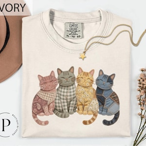 May include: Ivory-colored t-shirt featuring a graphic of four smiling cats with patchwork designs. The cats are in shades of pink, gray, yellow, and blue. A gold necklace with a star pendant and a sprig of dried flowers are also visible.