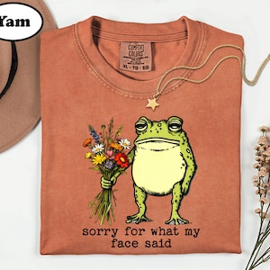 May include: A terracotta-colored t-shirt featuring a graphic of a green frog holding a colorful bouquet of wildflowers. Below the frog, the text reads, "sorry for what my face said." The shirt has a Comfort Colors brand label at the neck.