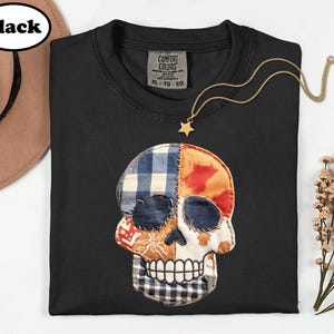 May include: A black t-shirt featuring a large, central skull graphic composed of various fabric patches in blue gingham, orange, and brown patterns. The skull is outlined with visible stitching. The shirt has a Comfort Colors label in size XL.