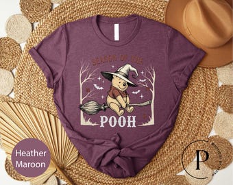 May include: A heather maroon t-shirt featuring a graphic of a cartoon bear wearing a witch hat and riding a broomstick. The text above reads Season of the and below reads Pooh. The design includes silhouettes of bare trees, small bats, and stars.