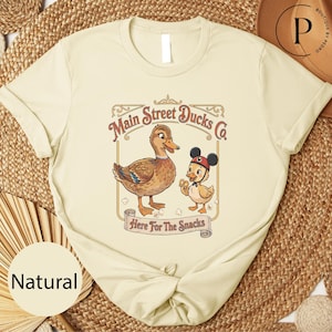 A beige t-shirt with a graphic design featuring two cartoon ducks, one larger and one smaller, along with the text "Main Street Ducks Co." and "Here for the Snacks".