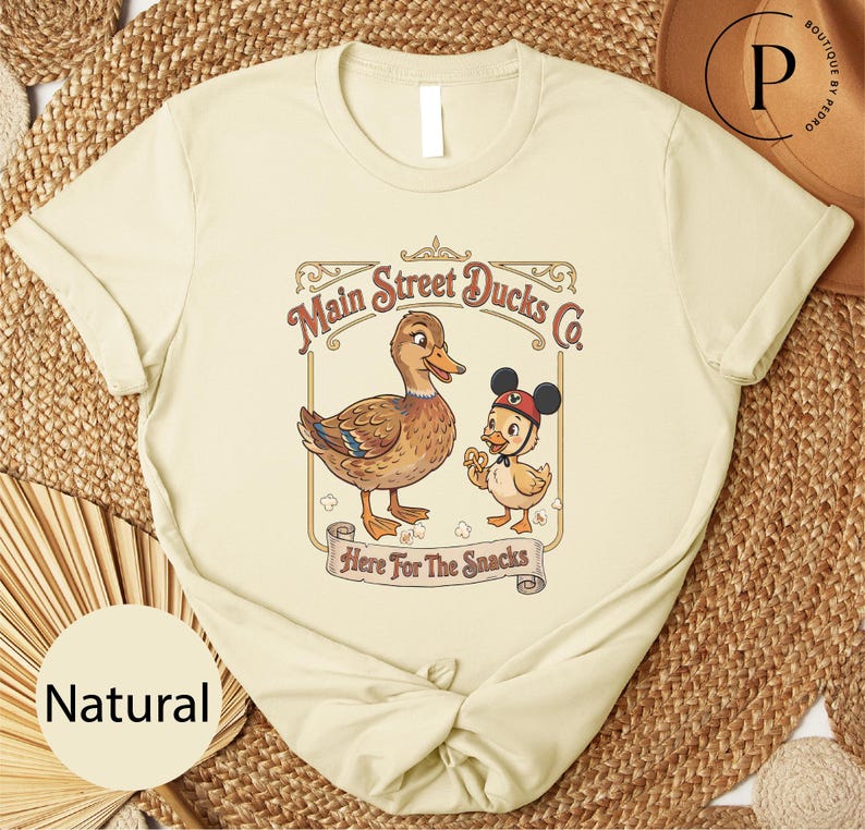 A beige t-shirt with a graphic design featuring two cartoon ducks, one larger and one smaller, along with the text "Main Street Ducks Co." and "Here for the Snacks".