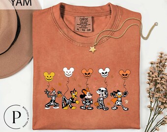 May include: A rust-colored crew neck t-shirt featuring a graphic print of five cartoon characters wearing skeleton costumes. Each character holds a string attached to a floating jack-o'-lantern or skull balloon. The shirt has a Comfort Colors label.