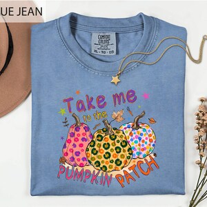 A blue t-shirt with a pumpkin patch design and the text "Take me to the pumpkin patch" printed on it, along with a necklace and a hat.