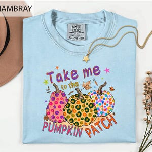 a light blue t-shirt with a colorful graphic design of three spotted pumpkins and the text "Take me to the pumpkin patch" printed on it.