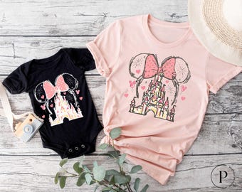 May include: A pink and black t-shirt set featuring a pink bow and a sketch of Cinderella's castle with fireworks. The shirts have a Mickey Mouse ear design.