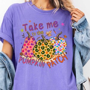 A woman wearing a purple t-shirt with a graphic design of three colorful, spotted pumpkins and the text "Take me to the pumpkin patch" printed on it.