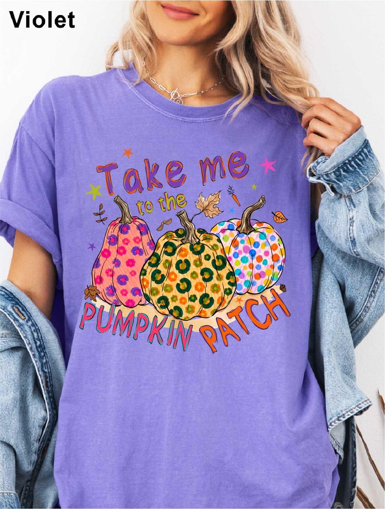 A woman wearing a purple t-shirt with a graphic design of three colorful, spotted pumpkins and the text "Take me to the pumpkin patch" printed on it.