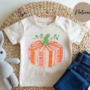 A white t-shirt with a graphic of a pumpkin and books, accompanied by a crocheted stuffed animal and a pair of blue jeans, all placed on a woven placemat.