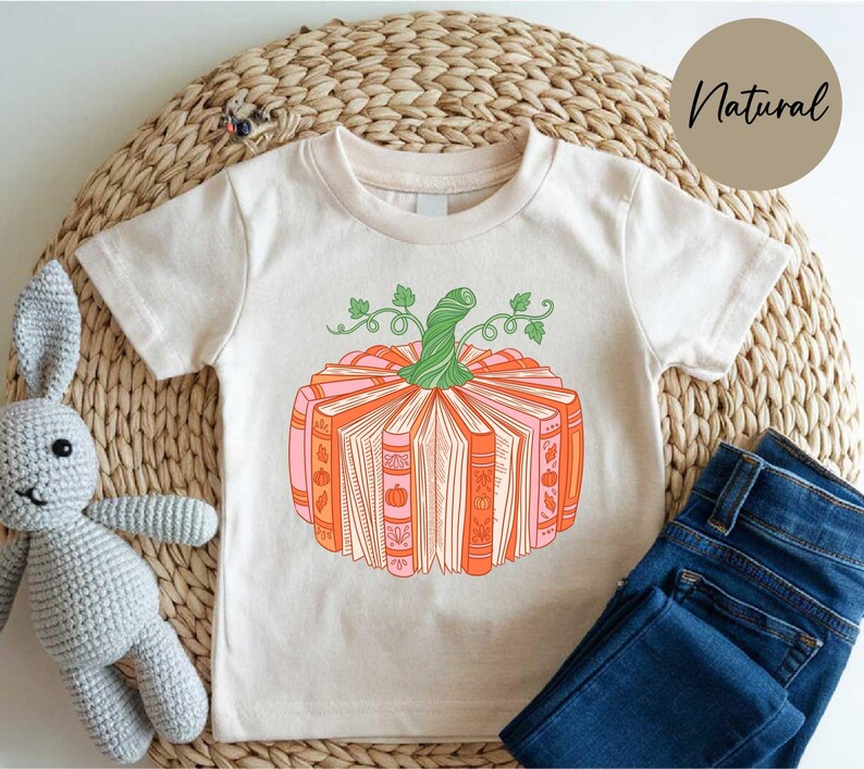 A white t-shirt with a graphic of a pumpkin and books, accompanied by a crocheted stuffed animal and a pair of blue jeans, all placed on a woven placemat.