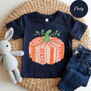 A navy blue t-shirt with a pumpkin graphic and a crocheted gray rabbit toy, along with a pair of blue jeans, are arranged on a woven placemat.