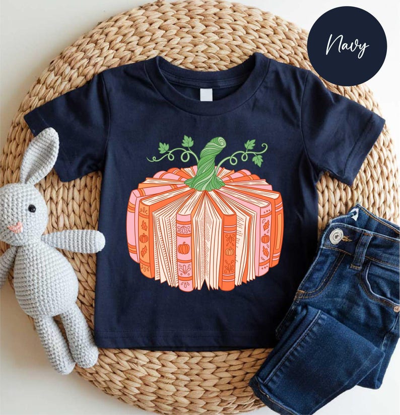 A navy blue t-shirt with a pumpkin graphic and a crocheted gray rabbit toy, along with a pair of blue jeans, are arranged on a woven placemat.