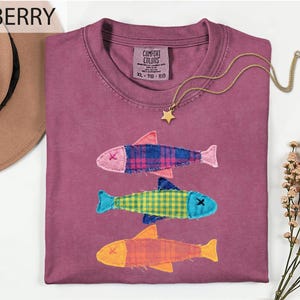 May include: A berry-colored crew neck t-shirt featuring three fish appliqués made from plaid fabric in pink, blue, yellow, and orange. The shirt has a Comfort Colors label at the neck and is displayed with a gold star necklace and a tan hat.
