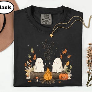A black t-shirt with a Halloween-themed graphic design, including two ghostly figures, a campfire, pumpkins, and autumn leaves.