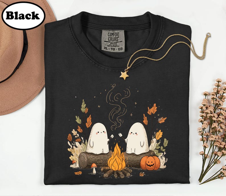 A black t-shirt with a Halloween-themed graphic design, including two ghostly figures, a campfire, pumpkins, and autumn leaves.