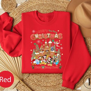 A red Christmas-themed sweatshirt with a cartoon image of Disney characters is displayed on a woven mat, along with a brown hat and a red fan.
